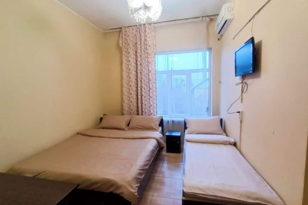 A room with two single beds, a small table, a television mounted on the wall, and a window with curtains.