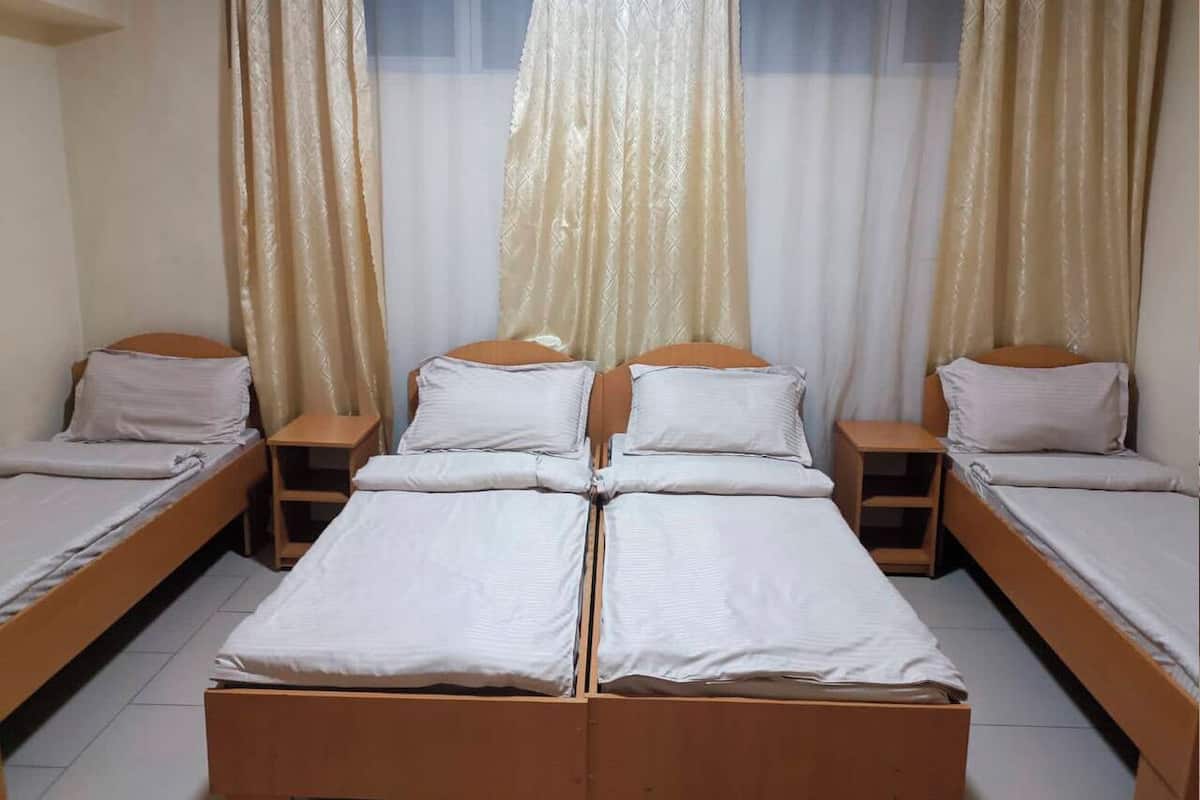 A room with two single beds and a double bed, each with wooden frames and white bedding. There are two nightstands with lamps, and the room has a window with sheer curtains.
