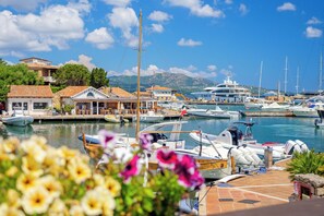 Un port de plaisance où sont amarrés différents types de bateaux, un restaurant appelé « Piratti » avec une terrasse, et un paysage montagneux en arrière-plan.