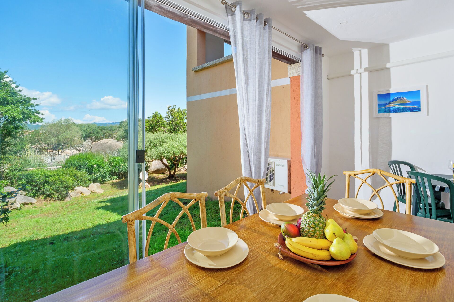 A dining table with chairs, plates, and a fruit bowl with bananas, pineapples, and pears.