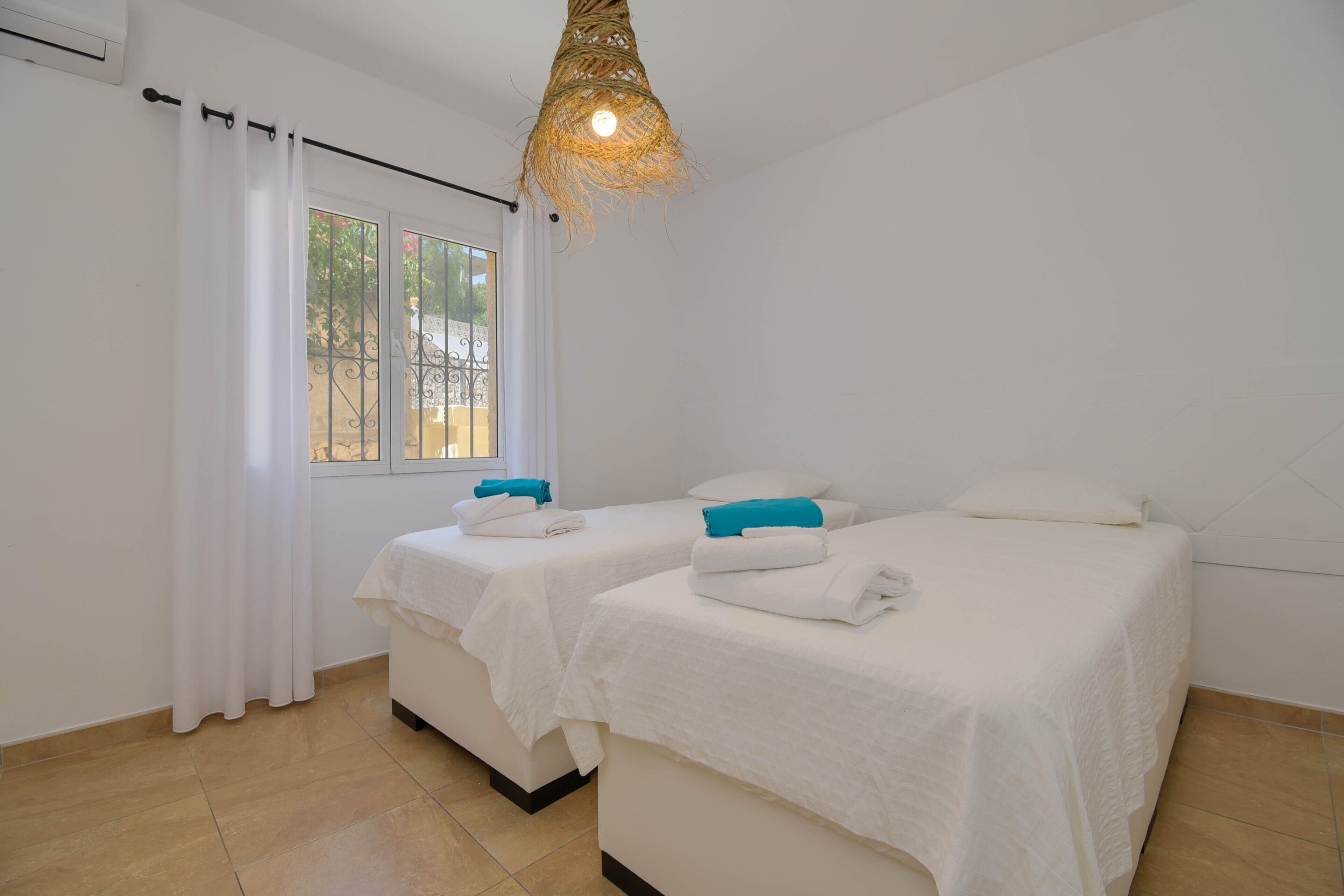A room with two beds, white bedding, a window with curtains, and a hanging light fixture.