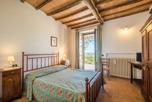 A bedroom with a bed, bedside tables, a radiator, a chair, and a window with curtains. - Podere Giardino Wine & Bed – Melograno Apartment: Country House with Pool, Wi-Fi & AC (Montalcino)
