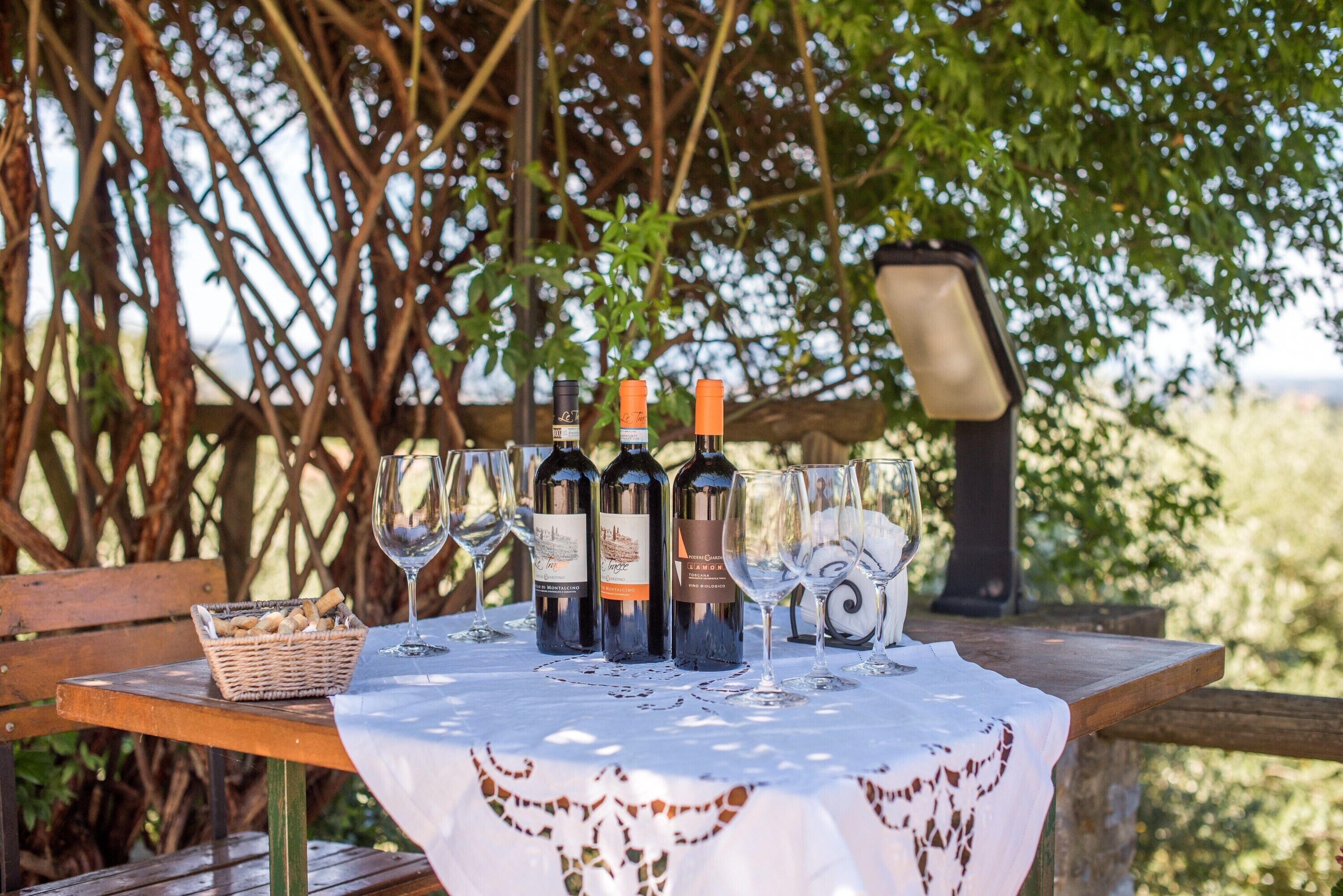 A table set outdoors with wine bottles and glasses, a basket of bread, and a wooden bench.