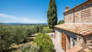 Un bâtiment en pierre avec un toit en tuiles, une porte en bois et une vue sur des oliviers et des collines lointaines.