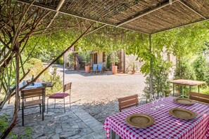 An outdoor patio with tables and chairs, a covered area with a thatched roof, and a view of a building in the background. - Podere Giardino Wine & Bed – Melograno Apartment: Country House with Pool, Wi-Fi & AC (Montalcino)