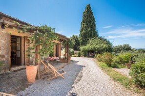 Un bâtiment rustique en pierre, avec une allée de gravier, une pergola en bois et un espace extérieur aménagé pour s’asseoir.