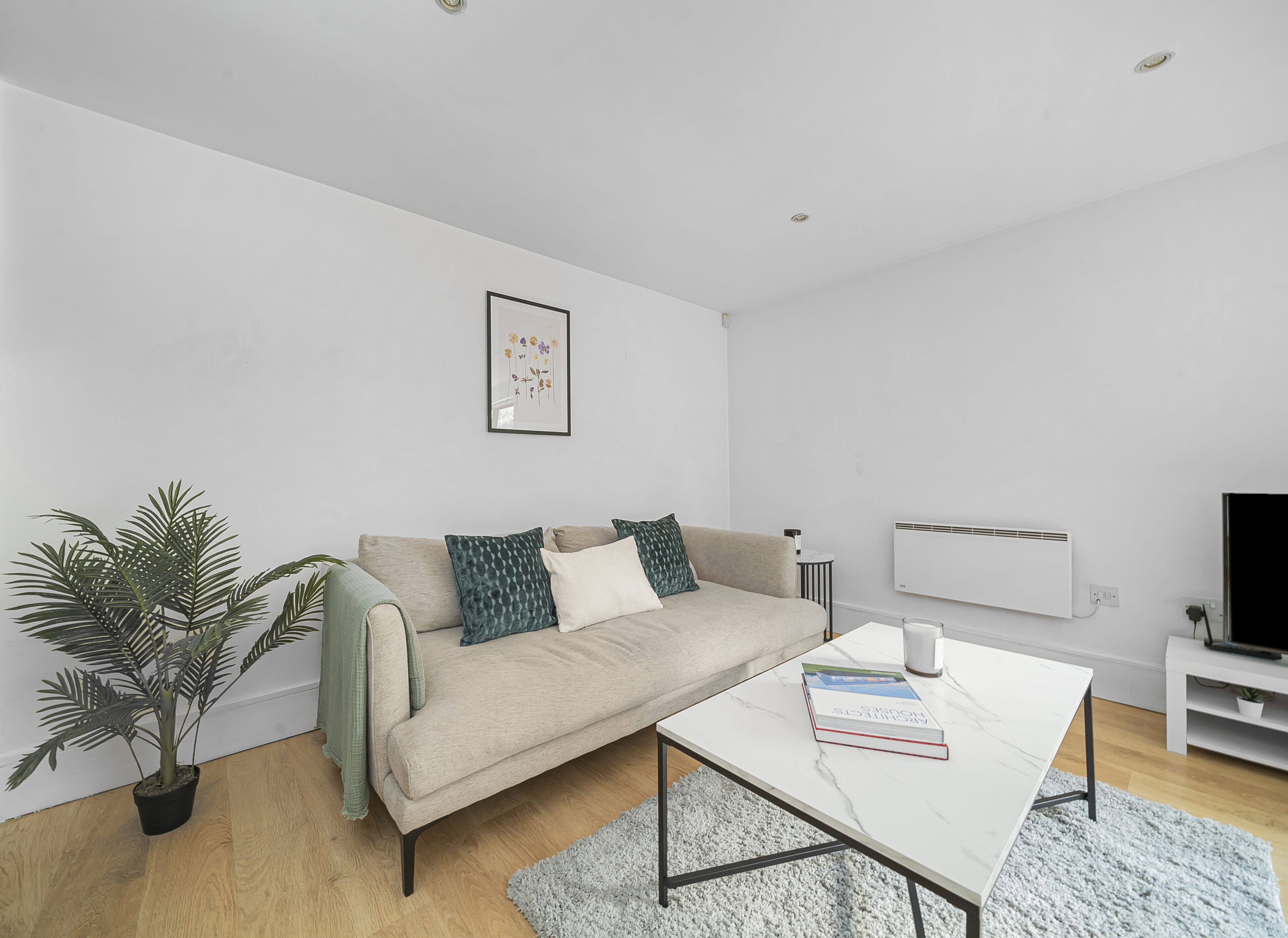 A modern living room with a beige sofa, a white coffee table, a framed floral artwork, a potted plant, and a white radiator.