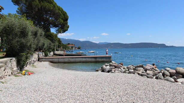 A pebbled beach with a dock, boats, and a rocky shoreline. - Holiday Apartment "Casetta - New Lake" with Private Terrace & Wi-Fi (Gardone Riviera)
