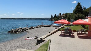 A waterfront patio with outdoor seating and umbrellas, a rocky shoreline, and a view of the water. - Holiday Apartment "Casetta - New Lake" with Private Terrace & Wi-Fi (Gardone Riviera)