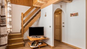 Una escalera de madera que lleva a una plataforma con un televisor sobre un soporte de madera, un estante pequeño con objetos, y una puerta con un cuadro en la pared.