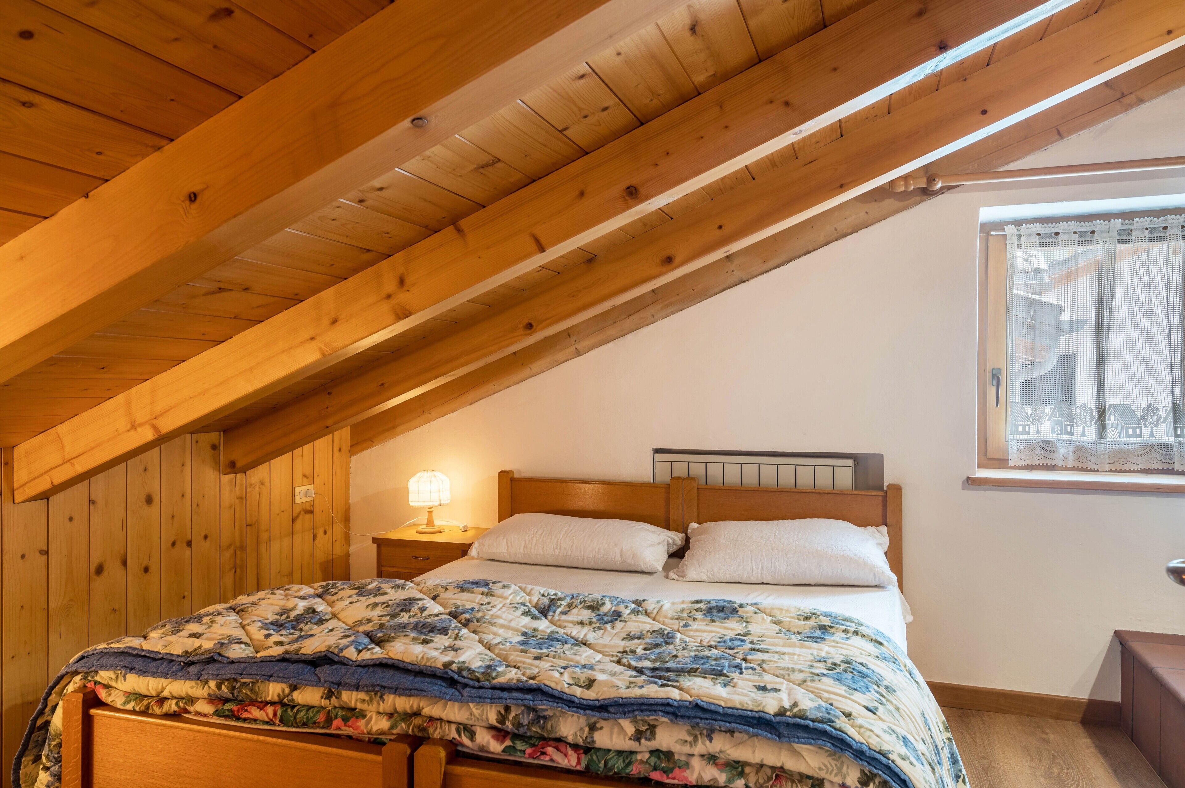 Un dormitorio con techo inclinado, una cama de madera, una mesita de noche con lámpara y una ventana con cortinas de encaje.