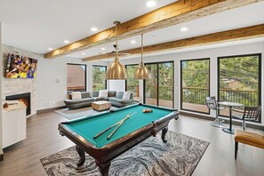 A modern living room with a pool table, comfortable seating, and large windows offering a view of the outdoors. - Luxury 6 bedroom retreat located right next to Bear Mountain! Central A/C! PET FRIENDLY!  Movie Theater! Pool Table! HOT TUB! (Big Bear Lake)