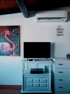 A room with a flat-screen TV on a white cabinet, a painting of a flamingo, an air conditioning unit, and a wall-mounted sign with the word 'love'.