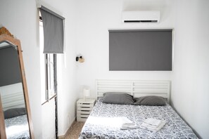 Ein Schlafzimmer mit einem Bett, einem Fenster mit Vorhang, einem Nachttisch mit Lampe und einer Klimaanlage an der Wand.