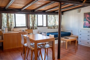 Une pièce de taille modeste, avec une table et des chaises en bois, un lit, un bureau et une fenêtre avec des rideaux.