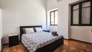 A bedroom with a bed, a window with blinds, a nightstand, and an air conditioning unit. - Apartment "Casa na Vila T2" with Shared Terrace & Wi-Fi (Fuseta)