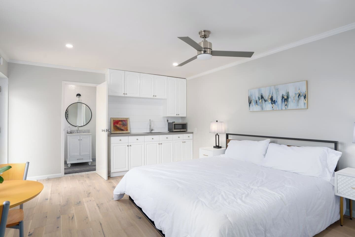 A bedroom with a bed, a ceiling fan, a painting, and a kitchen area with cabinets and a microwave.