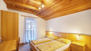 Un dormitorio con piso de madera, un escritorio, dos camas con ropa de cama floral, una ventana con cortinas de encaje y techo de madera.