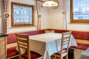 Une pièce comprenant une table, des chaises, un banc, une fenêtre avec des rideaux, une horloge et un luminaire fixé au mur.