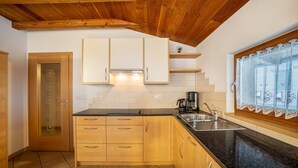 A kitchen with wooden cabinets, a black countertop, a window with lace curtains, and a tiled floor. - Apartment 'Speckign  3' with Mountain View, Balcony and Wi-Fi (Ahrntal)
