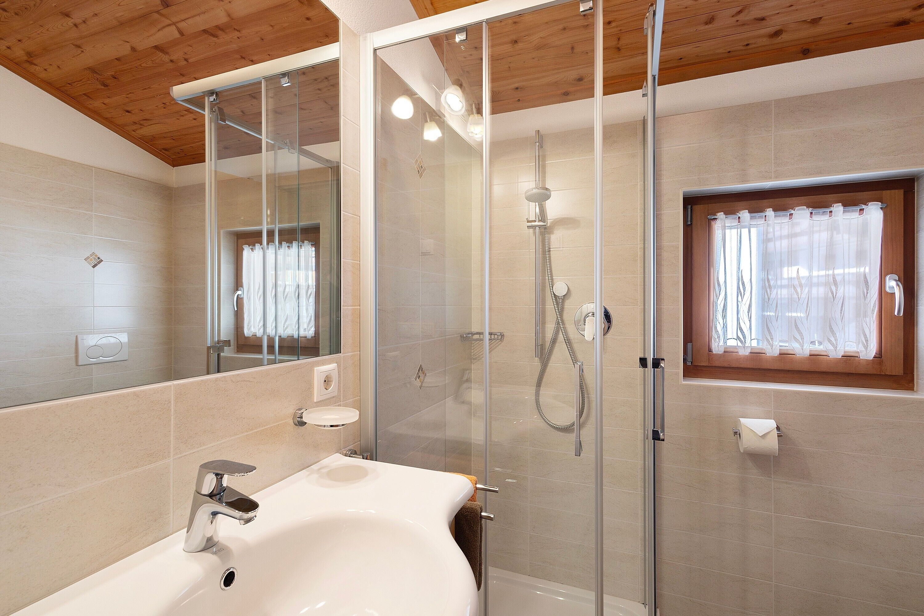 Ein modernes Badezimmer mit einer gläsernen Duschkabine, einem Waschbecken mit Spiegel und einem Fenster mit Vorhängen.