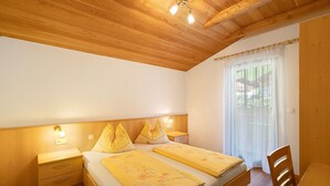 Un dormitorio con techo de madera, dos camas con ropa de cama floral, una mesita de noche, una silla y un ventanal con cortinas translúcidas.