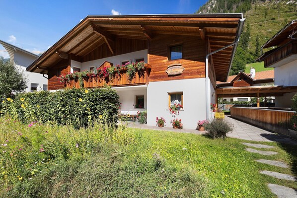 A wooden chalet with a balcony, surrounded by lush greenery and a clear sky.
