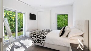 A modern bedroom with a bed, a flat-screen TV, and large windows with a view of greenery. - Villa "Pine Lodge" gated residence with heated private pool (Vilamoura)