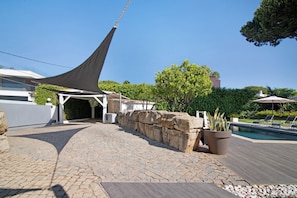 A patio area with a stone floor, a pool, and a pergola providing shade.