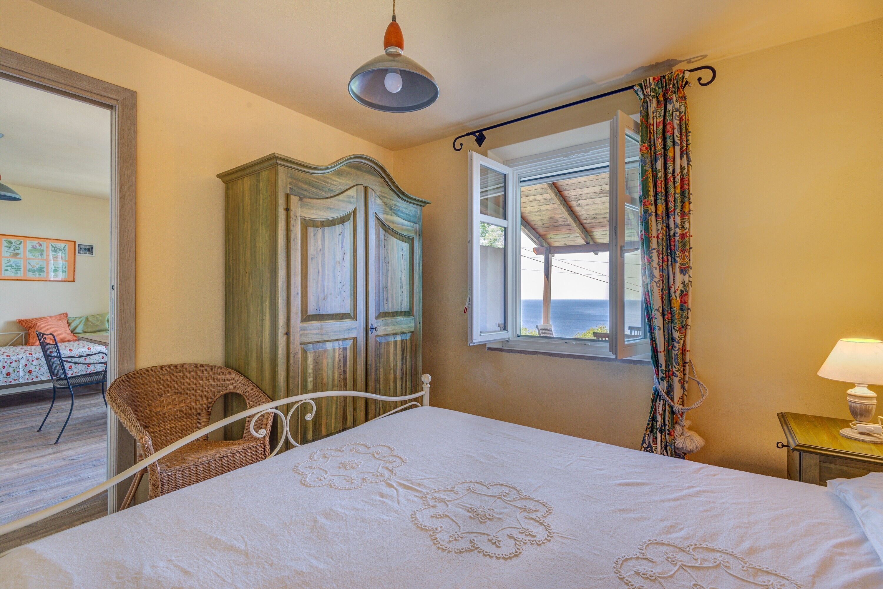 A bedroom with a bed, a wicker chair, a wardrobe, a window with curtains, and a lamp on a bedside table.