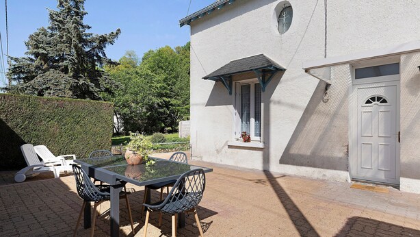 Un patio con una mesa y sillas de vidrio, una puerta blanca y un seto al fondo.