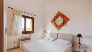Ein Schlafzimmer mit Bett, einem Fenster mit Vorhängen, einem wandmontierten Spiegel, einem Nachttisch mit Lampe und einer Heizung.