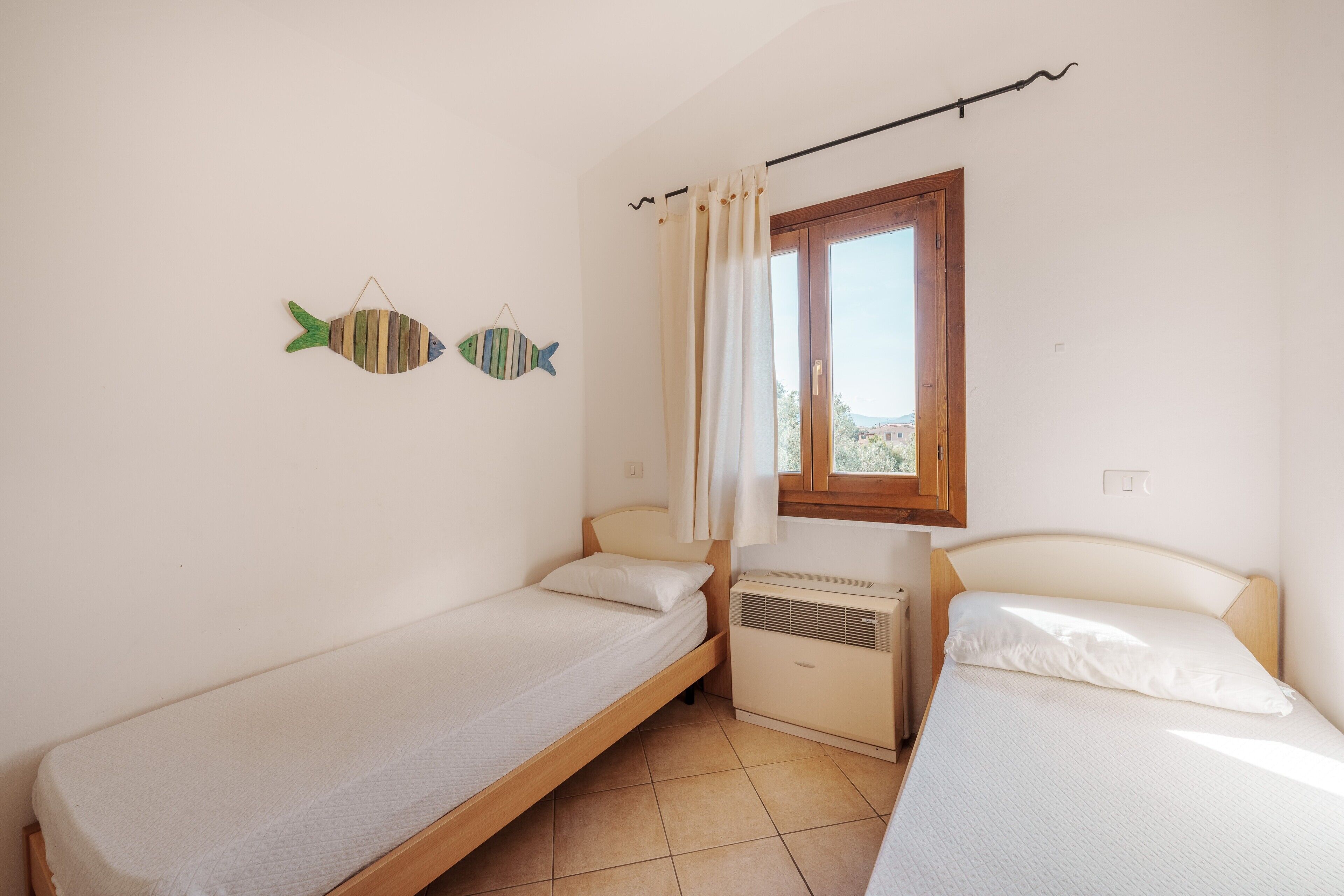 A small, clean room with two single beds, a window with curtains, and a wall-mounted air conditioner.