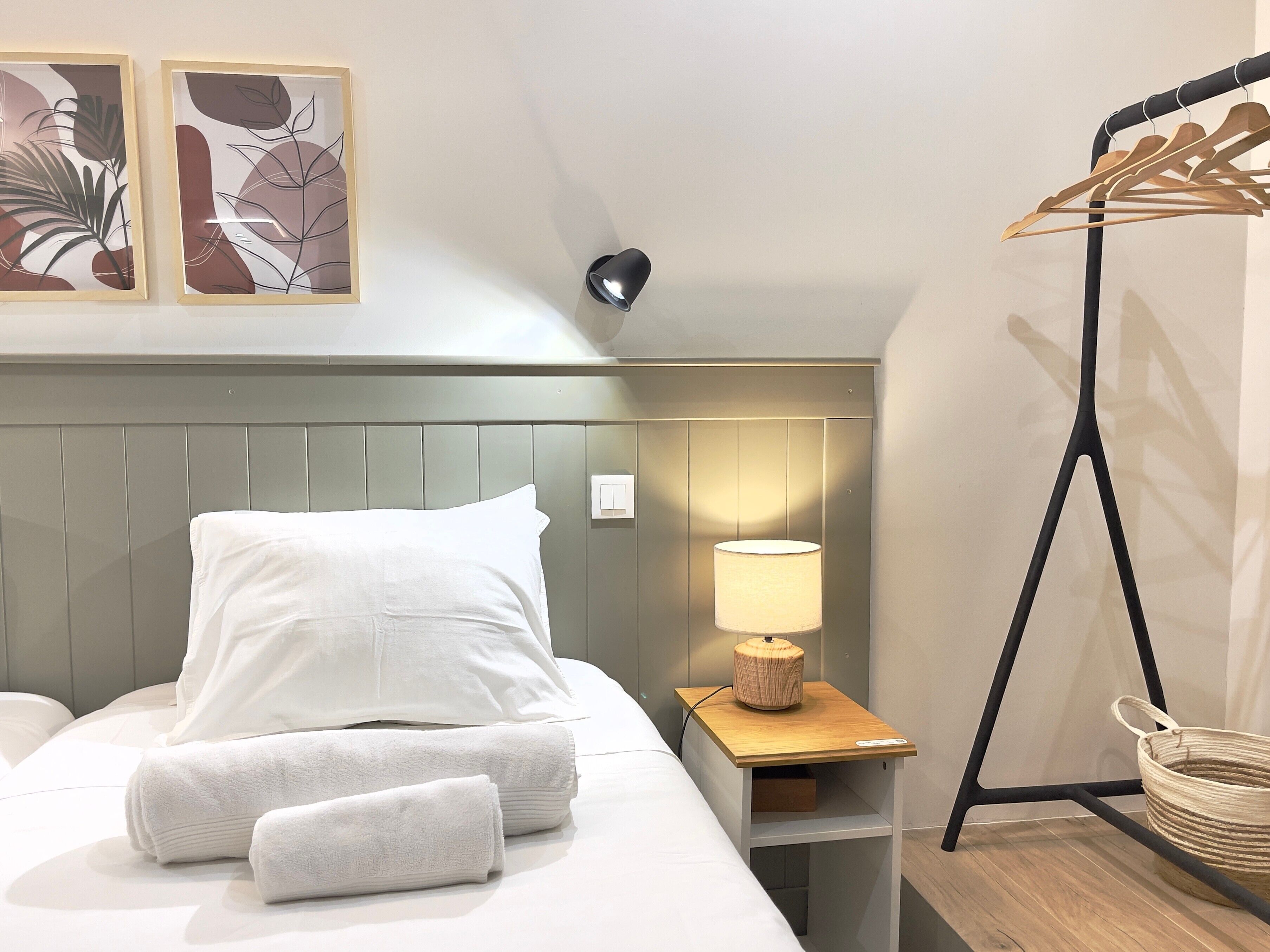 Una cama bien hecha con sábanas blancas, una lámpara de noche, una mesita de madera y un perchero de pared.