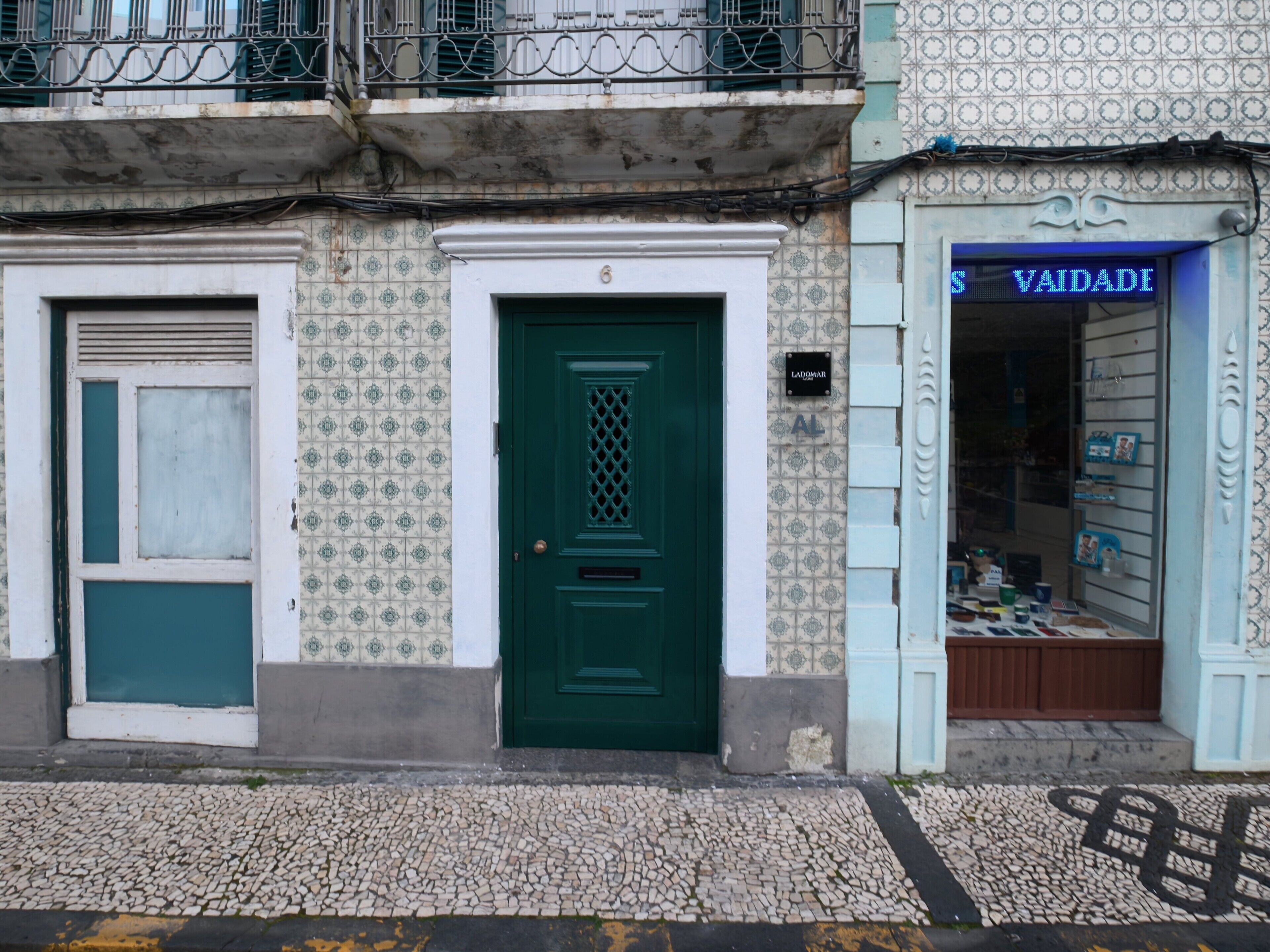 Un edificio con puerta verde, una tienda con cartel que dice 'VAIDADE' y una pared de azulejos con diseño decorativo.