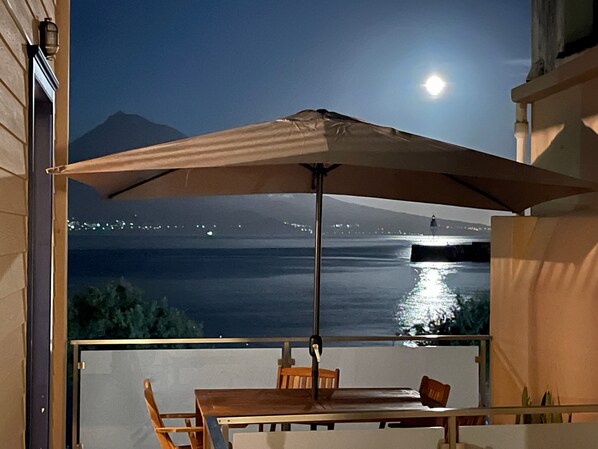 A balcony with a table and chairs, an umbrella, and a view of a body of water with lights and a mountain in the background.
