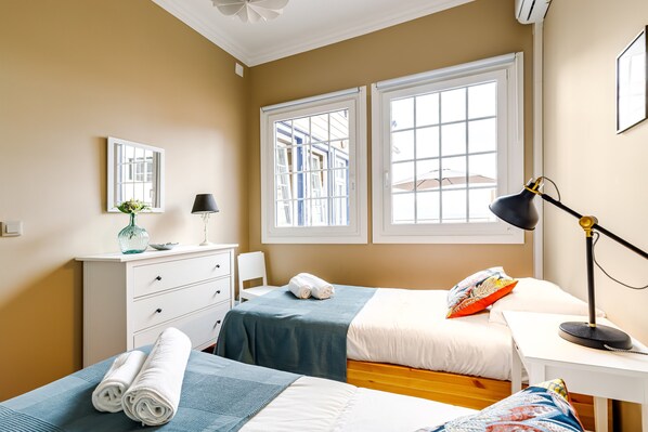 A bedroom with two beds, a desk, a lamp, a dresser, a mirror, and large windows.