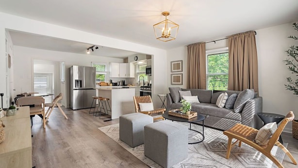 Ein modernes Wohnzimmer mit einem grauen Sofa, Poufs und einem Couchtisch. Im Hintergrund ist eine Küche mit Edelstahlgeräten zu sehen.