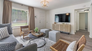 Ein modernes Wohnzimmer mit einer Couch, einem Hocker und einem Couchtisch. Ein Fernseher ist an der Wand montiert, darunter befindet sich ein Schrank.