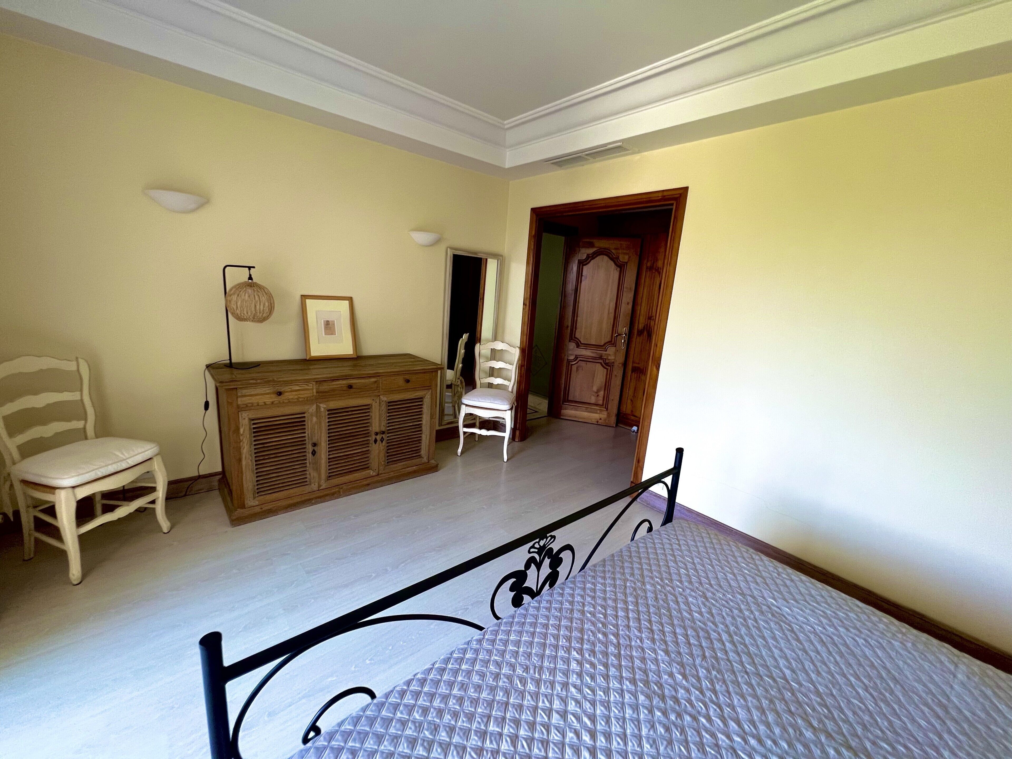 Une chambre à coucher comprenant un lit, une commode en bois, une chaise et une porte donnant sur une autre pièce.