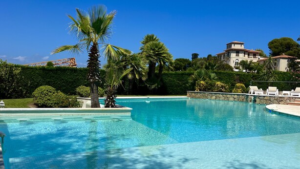 A clear blue swimming pool surrounded by palm trees and a stone wall, with a view of a building in the background. - Cap d'Antibes - Villa 10 people - Swimming pool (Antibes)