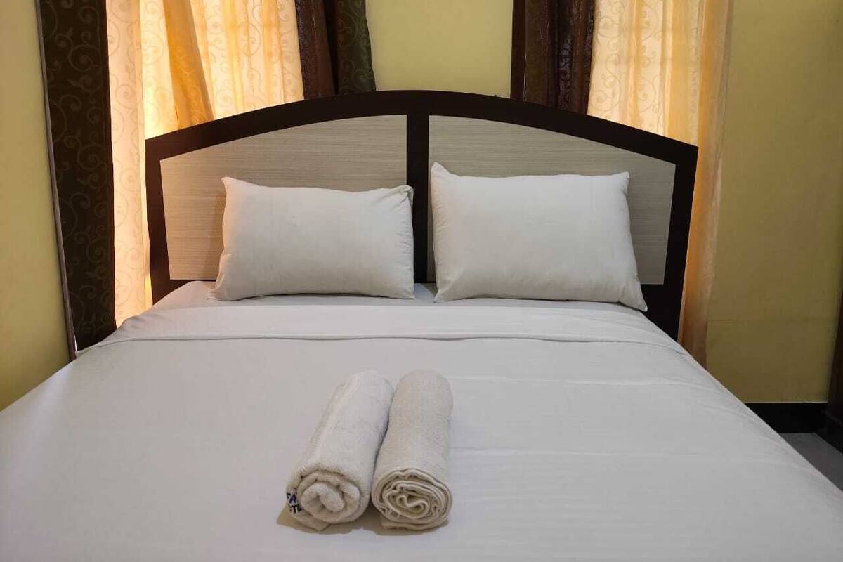 A neatly made bed with two pillows and rolled towels, set against a backdrop of patterned curtains.