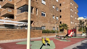 A playground with a seesaw and a slide, surrounded by a brick building with multiple balconies. - ACV - Torremar-2ª Linea Planta 5 Frontal 2 (OROPESA)