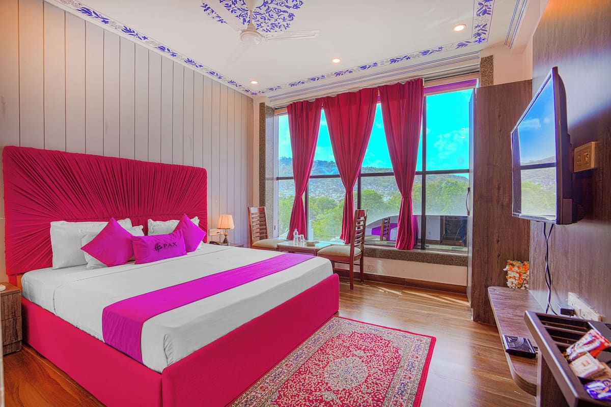 Una cama grande con cabecera roja, piso de madera, televisor y ventana con cortinas.