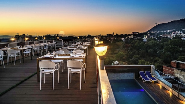 Una terraza en la azotea con mesas y sillas, una piscina y vistas a la ciudad al atardecer.