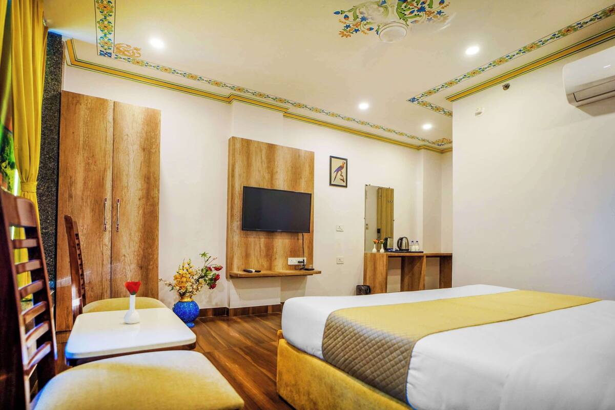 Habitación de hotel con dos camas, un televisor montado en la pared, una mesita con un florero y un armario de madera.