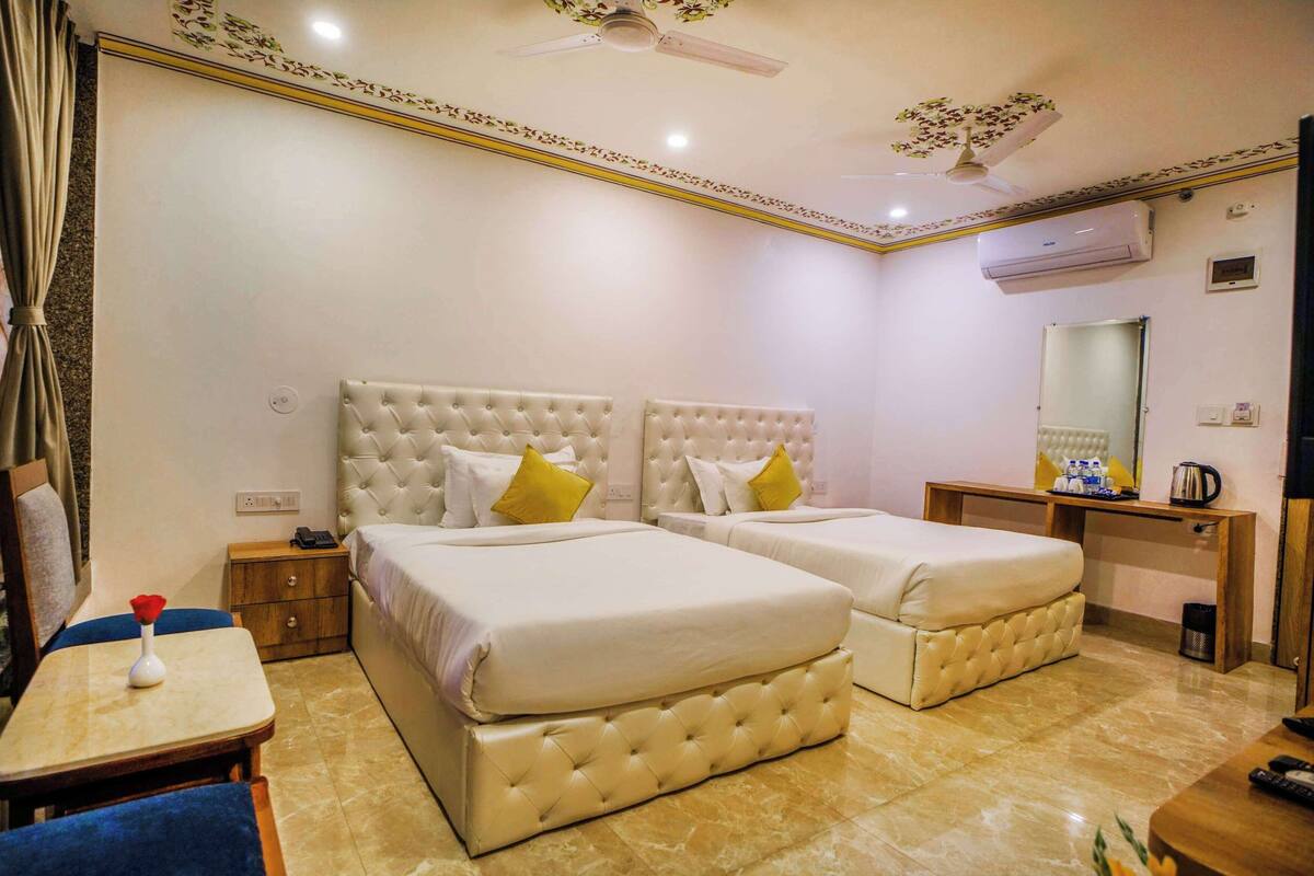 Habitación de hotel con dos camas, un escritorio, una silla, una mesita, una mesita de noche, un equipo de aire acondicionado, un espejo y un ventilador de techo.