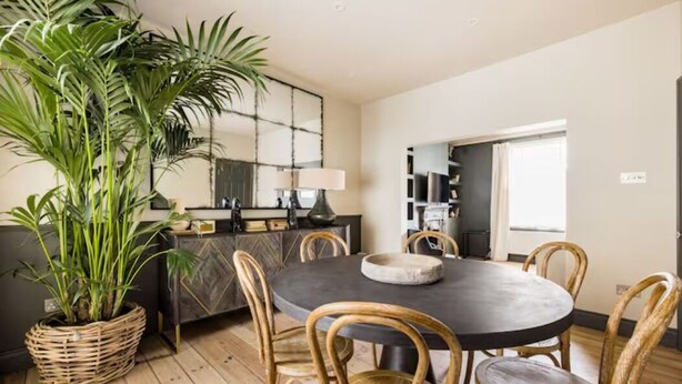 A dining area with a round table, six chairs, a large potted plant, a decorative mirror, and a modern kitchen in the background. - The North Kensington Cottage No.2 (London)