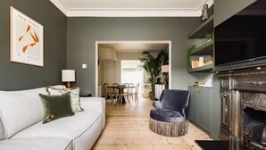 A living room with a white sofa, a leather armchair, a fireplace, and a dining area in the background. - The North Kensington Cottage No.2 (London)
