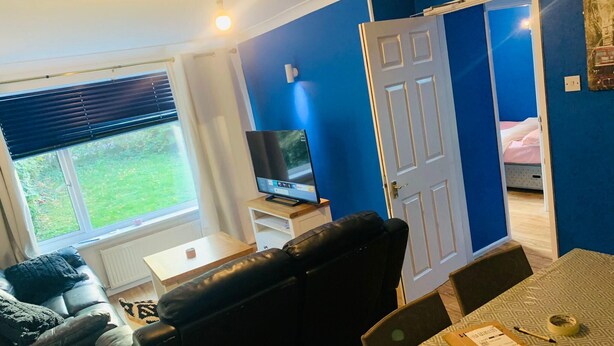 Un salon avec un canapé en cuir noir, un téléviseur posé sur un meuble, une table à manger avec des chaises et une porte donnant sur une chambre.